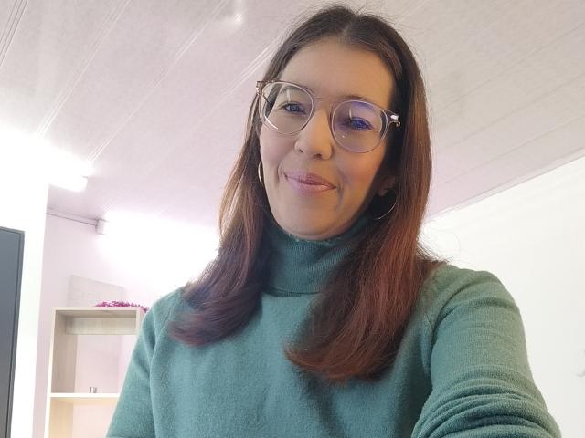 AntonnellaS with glasses, brown hair, green turtleneck, slight smile, medium-sized tits, white ceiling, white wall, shelf with pink flowers, subtle makeup, confident expression.