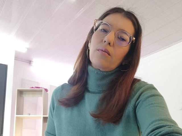 AntonnellaS with brown hair, glasses, green turtleneck, and hoop earrings, looking slightly to the side, in a bright room. Boobs not visible. Soft expression on her face.