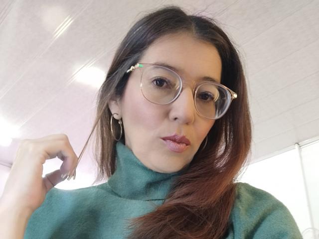 AntonnellaS with puffy pink lips, green turtleneck, hoop earrings, and big round glasses, holding brown hair, looking at camera.