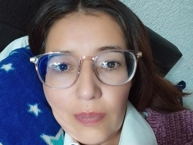 AntonnellaS has puffy pink lips, wearing clear glasses, with brown hair, lying on a blue and white blanket, and a white pillow, against a textured white wall.