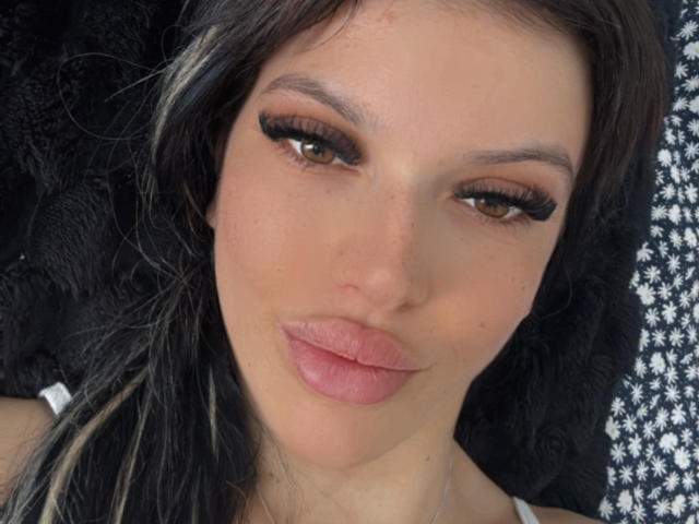 AndreeaG has brown eyes with thick, long eyelashes, pale pink lipstick, and black hair with blonde streaks. She's looking at the camera with a slight, pouty smile.