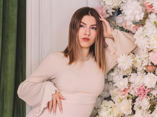 MaryBess with medium tits, wearing a beige dress, brown hair, pearl necklace, hand in hair, standing in front of a flower wall, green curtain on the left.