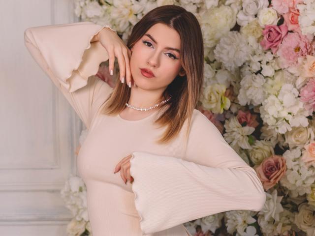 MaryBess with medium tits, wearing a sheer, ruffled, beige dress and pearl necklace, posing with one hand on her cheek, brown hair, pink roses, and white flowers in background.
