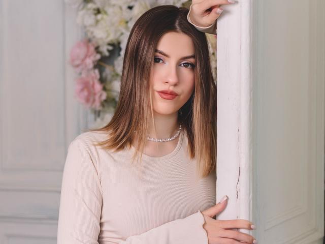 MaryBess with straight brown hair, pink lips, and a pearl necklace, wearing a beige long-sleeve top, leaning on a white wall with a gentle smile.