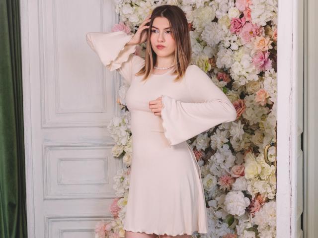 MaryBess with medium tits, wearing a pale pink dress with flared sleeves, standing against a floral wall, hand in hair, looking at camera, pearl necklace, green curtain.
