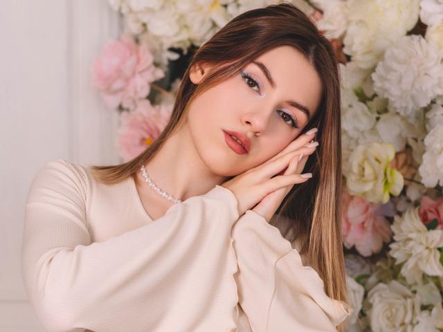 MaryBess with light brown hair, wearing a beige top, touching her face with manicured nails, pink lipstick, pearl necklace, soft makeup, white and pink flowers in the background.