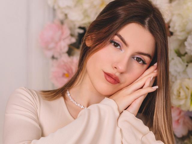 MaryBess has a pretty face, brown eyes, and light brown hair. She's wearing a beige top and a pearl necklace, with her hand on her cheek.