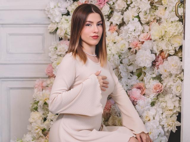 MaryBess with brown hair, wearing a beige dress, covering her tits with her hand, sitting in front of a floral wall, wearing a pearl necklace, looking at the camera.