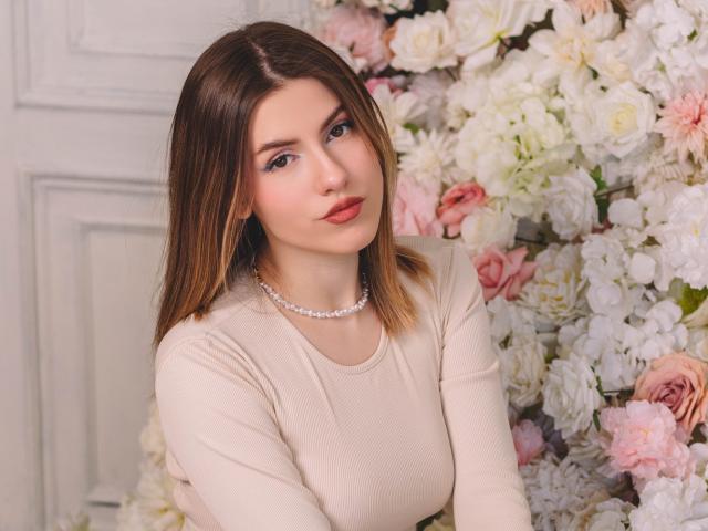 MaryBess with medium brown hair, red lipstick, and a pearl necklace, wearing a beige long-sleeve top, standing in front of a floral wall. Her tits are small.