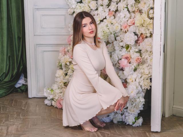 MaryBess, squatting, wearing a pale beige dress, pearl necklace, and clear heels, surrounded by white and pink flowers, has fair skin, brown hair, and a cute, sexy face.