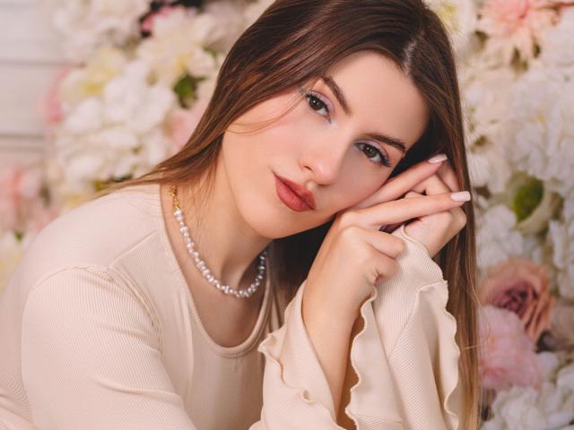 MaryBess with long brown hair, wearing a pearl necklace and beige top, resting her chin on her hand, showing off her pink lipstick and white nails.