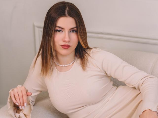 MaryBess with medium boobs, wearing a beige dress and pearl necklace, has straight brown hair, red lips, and is sitting on a white chair, looking at the camera.