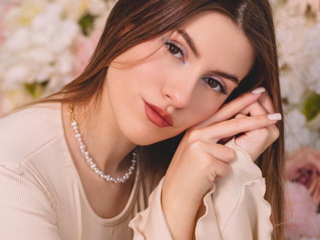 MaryBess has long brown hair, green eyes, and red lipstick, wearing a pearl necklace and beige top, with her hand near her face, showing off her white nails.