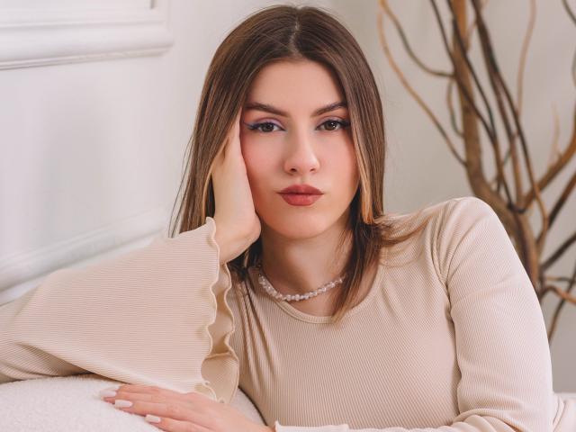 MaryBess with brown hair, wearing a beige ribbed top and pearl necklace, resting her cheek on her hand, looking at the camera with pink lipstick and subtle makeup.