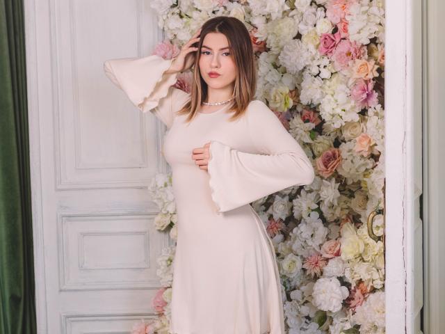 MaryBess, with tits covered by a white, long-sleeve dress, stands in front of a floral wall, hand in hair, looking at camera, wearing a pearl necklace.