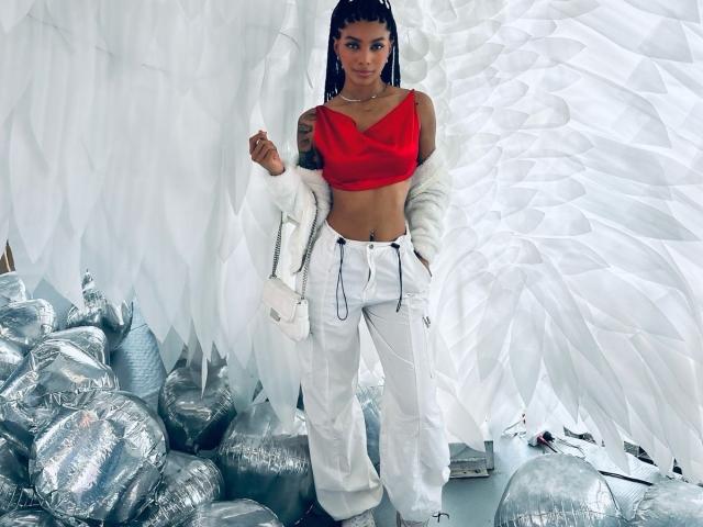 NaomiPortia with dark skin, red crop top, white fluffy jacket, white pants, hands in pockets, standing among silver balloons, looking at camera, long braids, tattoo on right arm.