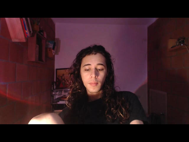 DussanHot69 with curly dark hair, black shirt, and pensive expression, sitting in dimly lit room, looking down at camera with a slight smirk, relaxed and natural.