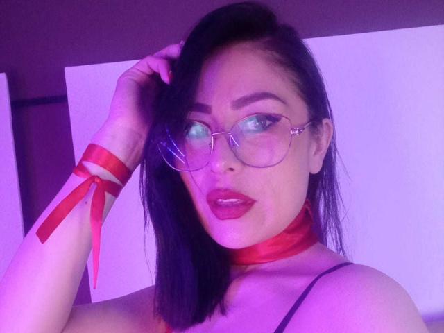 VioletaWhitee with dark hair, glasses, red ribbon choker, and matching red ribbon on wrist, looking at camera with red lipstick, purple and pink lighting.