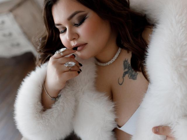 DreamForeYou with brown wavy hair, wearing a white fur coat, black nail polish, holding a ring, pearl necklace, tattoo on right shoulder, white bra, and looking down.