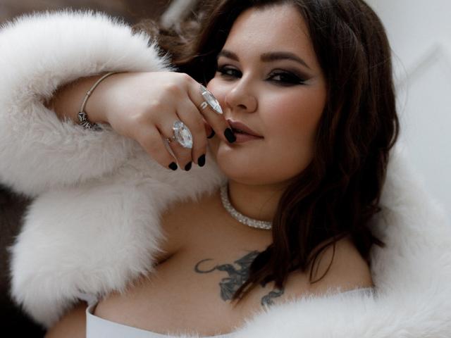 DreamForeYou with dark wavy hair, white fur coat, black nail polish, diamond rings, choker, and tattoo on chest, smirking, showing off her tits.