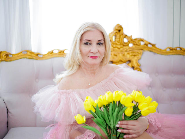 LadyJanyne with blonde hair, pink off-shoulder dress, holding yellow tulips, sitting on a gold and white tufted sofa, smiling, wearing a pearl necklace.