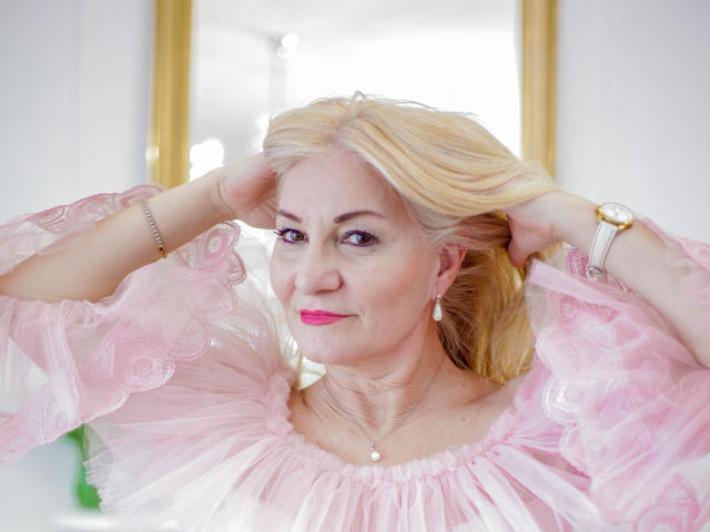 LadyJanyne with blonde hair, pink lace dress, pearl necklace, and pink lipstick, hands in hair, wearing a gold watch, smiling slightly, looking forward. Her tits are small.
