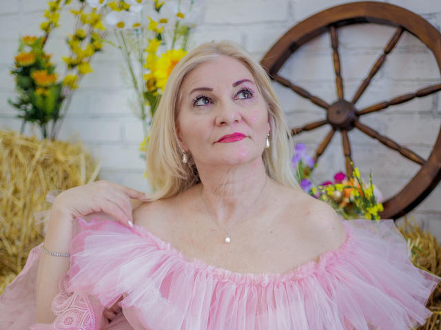 LadyJanyne with blonde hair, pink tulle dress, and pink lipstick, holding her dress, showing her tits, standing near a wooden wheel, with flowers and hay in background.