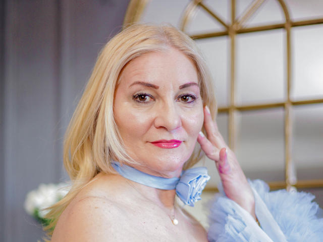 LadyJanyne with blonde hair, blue ribbon choker, pink lipstick, and light makeup, touching her cheek with a gentle smile, showing her bare shoulders and a light blue fur accessory.