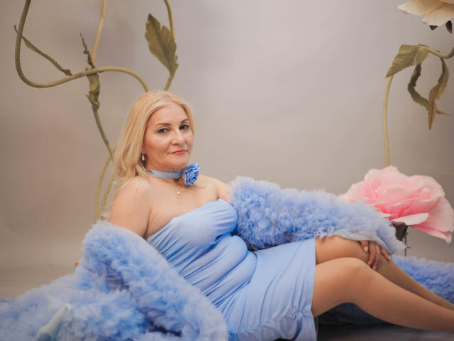 LadyJanyne with blonde hair, wearing a blue dress, blue choker, and fluffy blue robe, sitting with her legs crossed, showing her tits and a pink rose in the background.