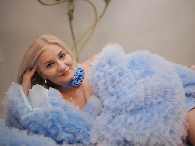 LadyJanyne with blonde hair, wearing a fluffy blue fur coat, blue flower on neck, white blouse, looking at camera, hand on head, soft smile, light makeup, earrings, calm expression.