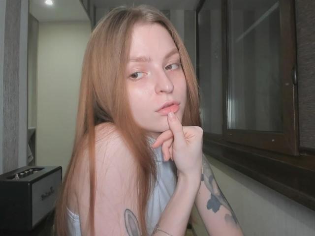 MalenaGolden, con lunghi capelli rossi, pelle chiara e tatuaggi sul braccio, che si morde un dito, guarda leggermente di lato con un'espressione sottile e sensuale..