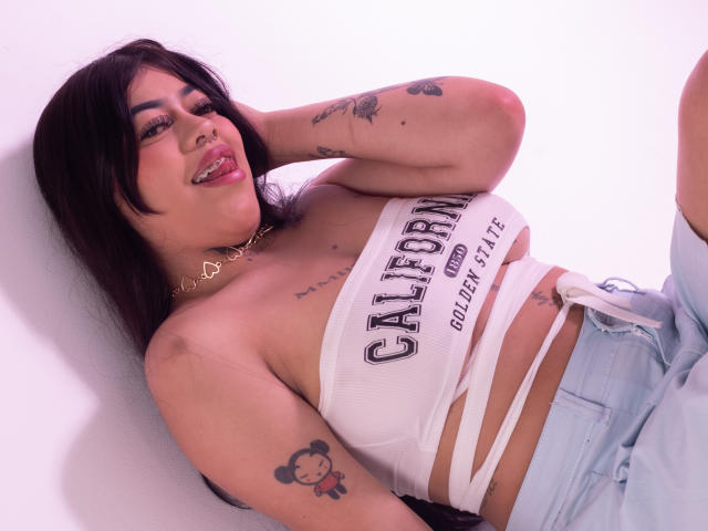 KarlySantana with black hair, tattoos, and a white tube top showing her tits, wearing light blue jeans, lying down, smiling, and looking at the camera.