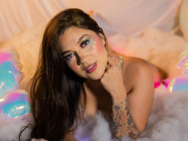 LindaJimenex with long brown hair, colorful eye makeup, tattoos on her right arm, lying in fluffy white foam, touching her neck, smiling with pink lips, and neon pink highlights.