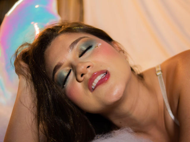 LindaJimenex with her eyes closed, makeup shimmering, lips parted, brown hair, white strap, and a sexy, foamy expression on her face. Her ass is partially visible.
