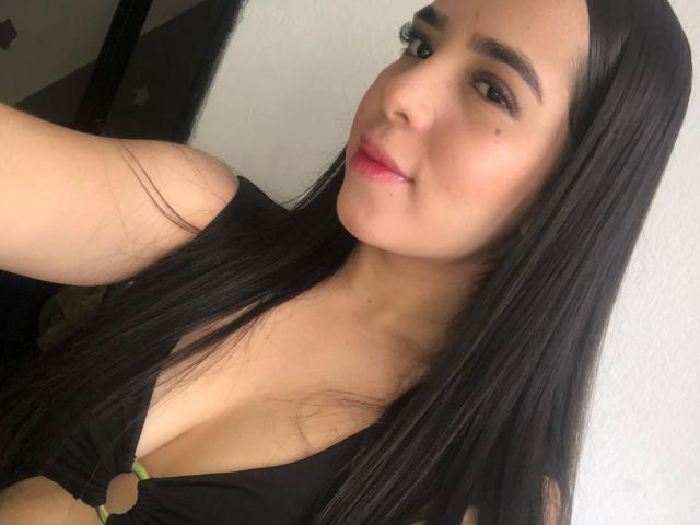 Alana Reyes, con cabello negro largo, vestida con una blusa negra con un anillo, mostrando sus senos, labios rosados y una pequeña peca en su mejilla derecha, sonriendo ligeramente..