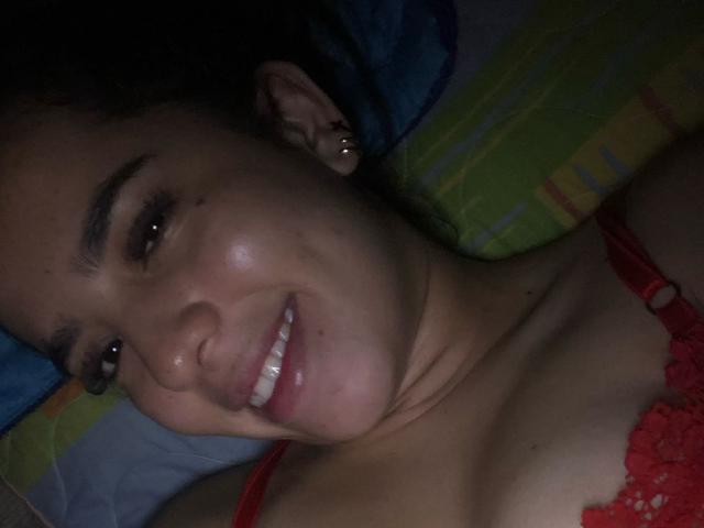 AlanaReyes with dark curly hair, red lace bra, smiling, showing white teeth, small mole above lip, ear piercings, lying down, dimly lit, slightly flushed, cute.