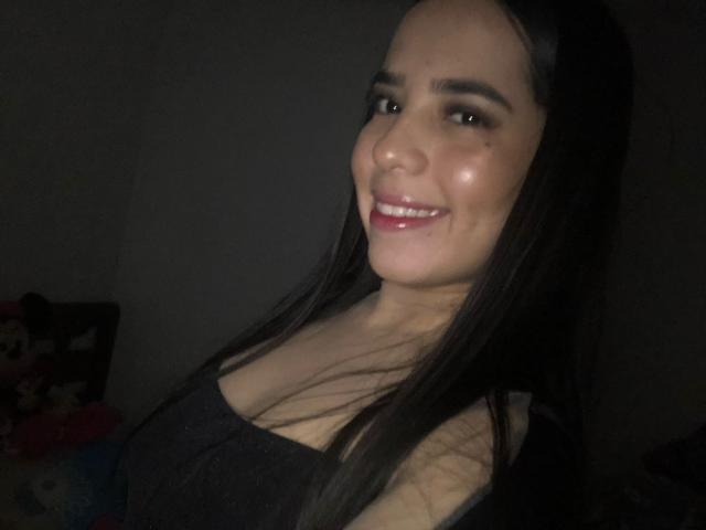 AlanaReyes met lang zwart haar, lachend met roze lippenstift, gekleed in een zwarte top, waarbij haar borsten zichtbaar zijn, in gedimd licht, kijkend naar de camera, donkere achtergrond, licht glanzende huid..
