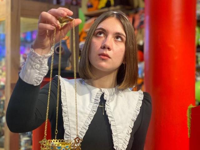 IsleBelle with shoulder-length brown hair, wearing a black dress with white ruffled collar, holding a golden chain with a small, ornate, red and gold bag.