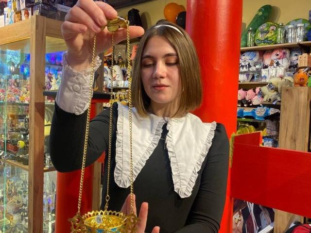 IsleBelle with short brown hair, wearing a black dress with white ruffled collar, holding a gold pendulum, looking down, standing in front of red columns, in a quirky shop.