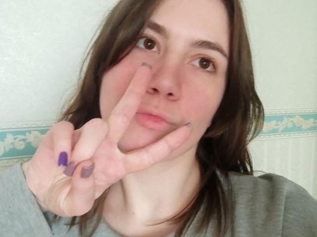 IsleBelle with brown hair, light skin, making a peace sign with purple and black nails, wearing a grey shirt, has a small tattoo near her right eyebrow.