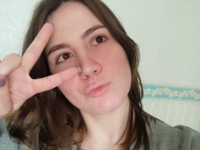 IsleBelle has a cute face, brown wavy hair, and is making a peace sign with her right hand, showing off her pinky and middle fingers. She's wearing a gray shirt.