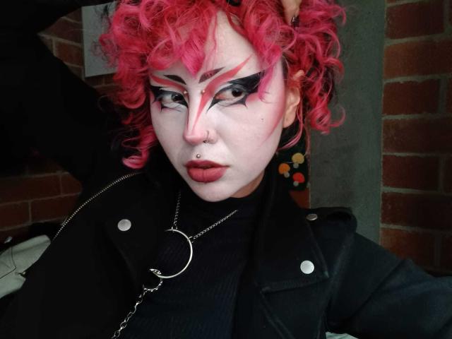GothVioleth met krullend roze haar, witte make-up, zwarte eyeliner, rode lipstick, een neusring, een zwarte jas en een metalen ketting. De borsten van GothVioleth zijn niet zichtbaar op deze foto..