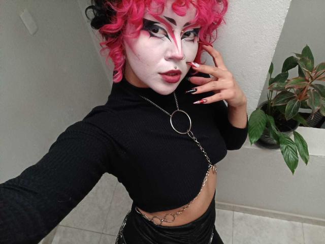 GothVioleth con capelli ricci rosa, trucco bianco sul viso, top nero, collana a catena e pantaloni in pelle nera, che mostrano una cintura a catena, una mano con le unghie rosse e uno sguardo sicuro..