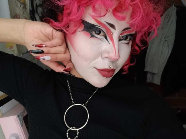 GothVioleth con cabello rizado rosa, maquillaje blanco, maquillaje de ojos negro y rojo, piercing en la nariz, top negro, collar con anillos y uñas rojo y negro, tocándose la oreja. El rostro de GothVioleth es pálido y el maquillaje es llamativo..