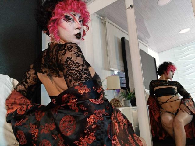 GothVioleth, avec ses cheveux roses bouclés, son haut en dentelle noire et sa robe à motifs rouge et noir, assise sur un lit, montrant son dos et son reflet dans un miroir, exposant des culottes noires à fines bretelles et son derrière nu..