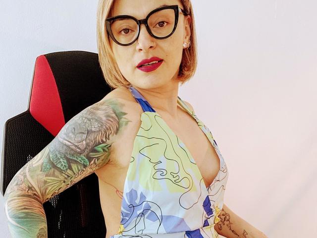 KatParadise com óculos, batom vermelho, cabelo curto loiro, blusa de alça azul estampada, mostrando os seios, braço tatuado, cadeira preta e vermelha, parede branca, sorrindo, selfie, parte superior do corpo..