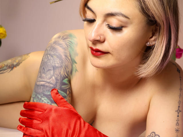 KatParadise with red gloves, red lipstick, and short blonde hair, looking down at her tattooed arm, showing a detailed lion tattoo, with visible tits and a nose ring.