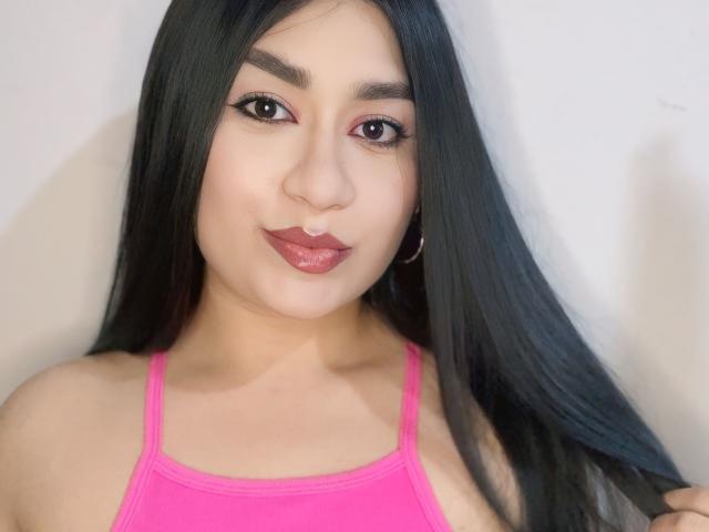 MiaDuboisX with long black hair, pink tank top, hoop earrings, and glossy red lips, looking at the camera with a slight smile. Her tits are small and cute.