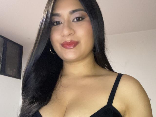 MiaDuboisX with long black hair, brown eyes, and red lipstick, wearing a black bra, showing her cleavage and small hoop earrings, smiling softly.