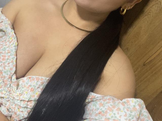 MiaDuboisX's tits are visible in a floral off-shoulder top, with a long black ponytail covering part of her chest. She's wearing a gold hoop earring and a thin necklace.